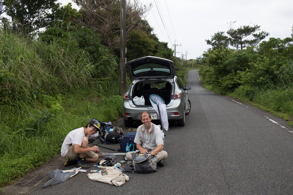 photo of two people sitting on the road with insect collecting gear