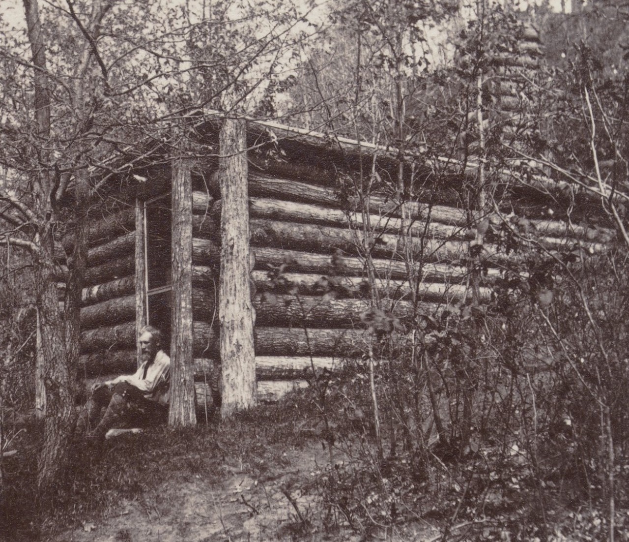 Black and white photo of a man sitting on the doorstep of a log cabin in the woods.