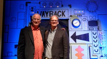 Two men in suit jackets and glasses smile and pose for a picture in front of a prop time machine