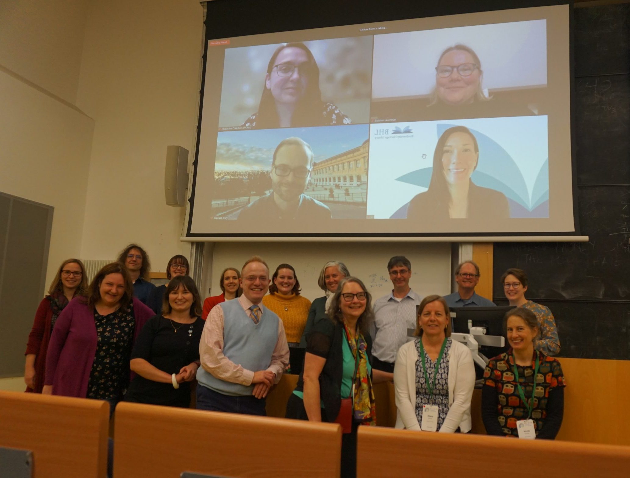 A group of conference attendees pose for a group photo in front of a projection screen with virtual attendees on screen