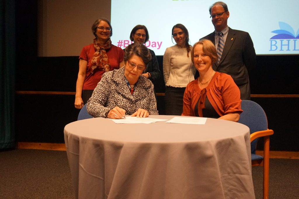 Nancy Gwinn signs the MOU for BHL Australia while colleagues pose for a group photo in front of a large screen