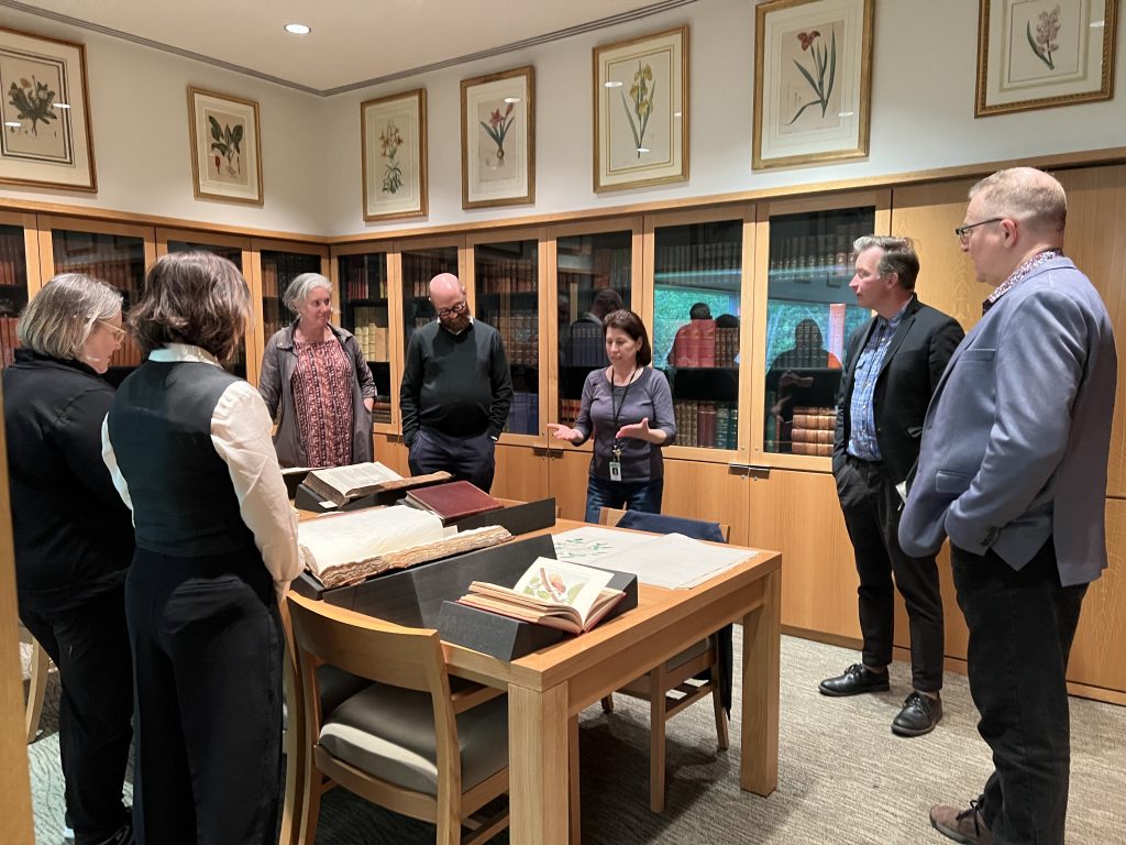 A group of people listen to a woman standing in front of rare books on book cradles in a room full of locked book cabinets and botanical art