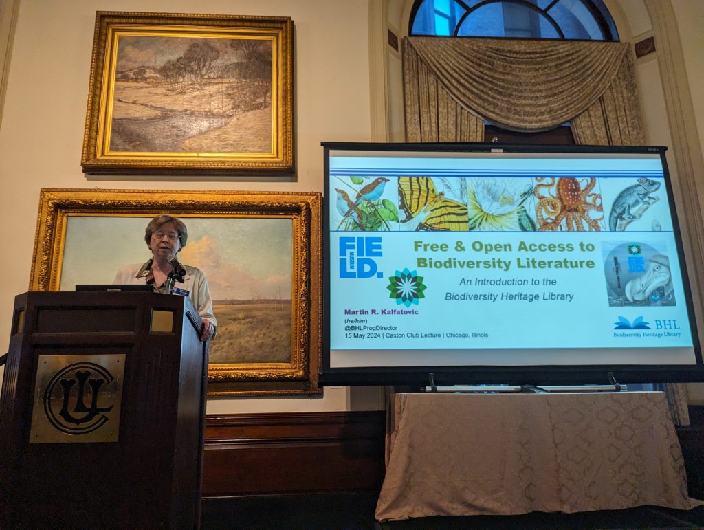 A woman with short hair and glasses speaks at a podium with a display screen behind her. The screen displays a slide with biodiversity images and the title Free and Open Access to Biodiversity Literature - an introduction to the biodiversity heritage library