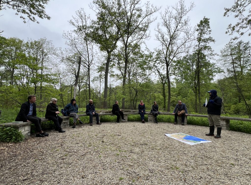 A man presents a map to a group sitting in a half circle of wooden benches in a clearing in the woods