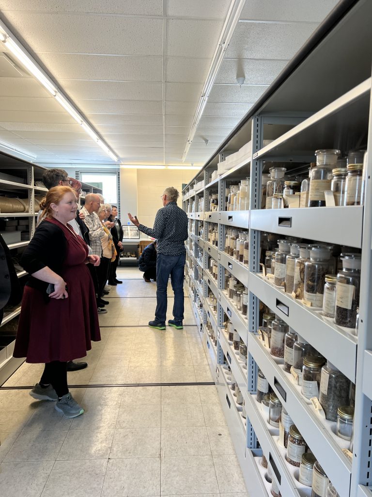 A group of people listen a man describes a collection of botanical samples in glass jars on a storage shelf