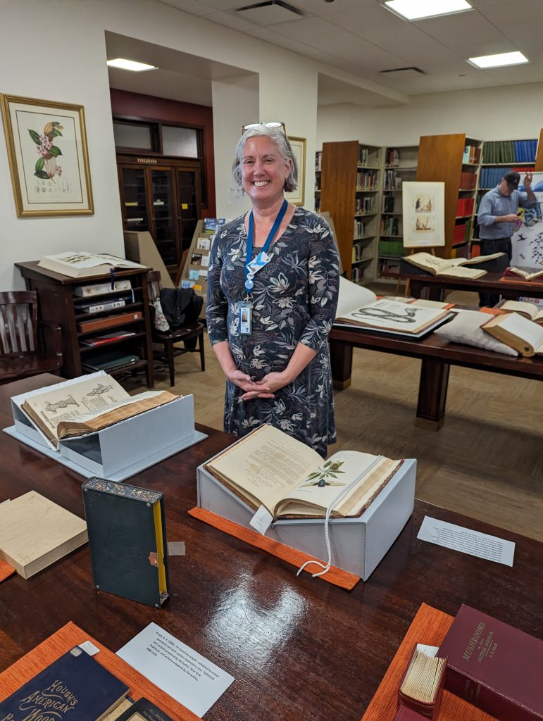 A woman smiles and poses behind rare books on display on a wooden table