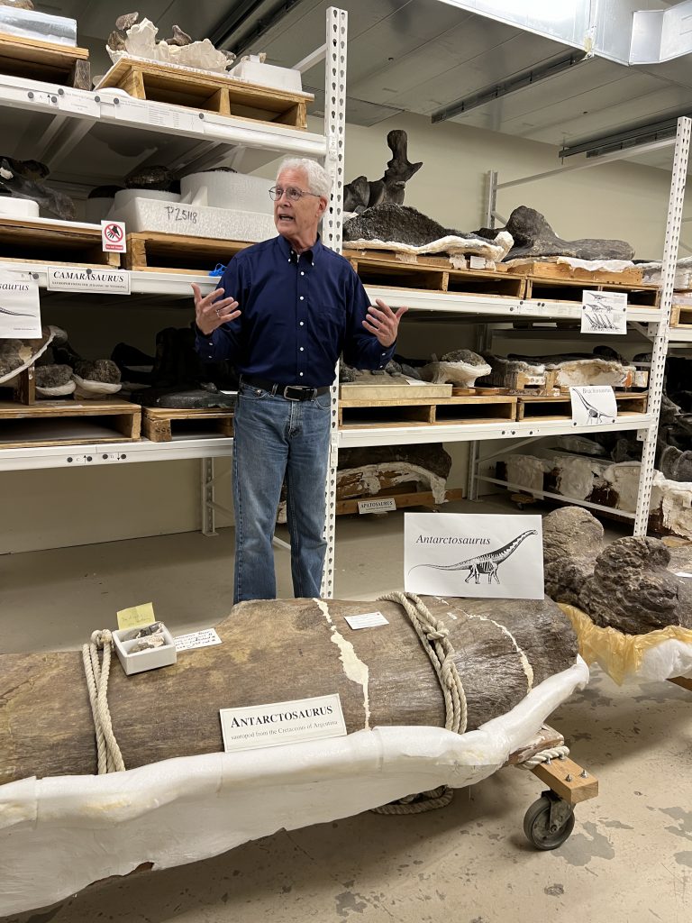 A man gestures while talking in a room full of dinosaur fossils