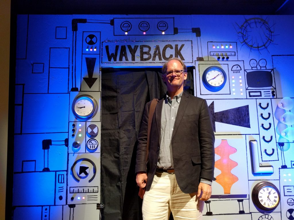 A man with glasses, wearing khaki pants and blazer, stands in front of a cardboard machine labeled "Wayback"