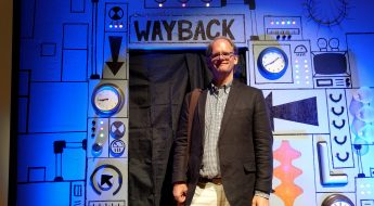 A man with glasses, wearing khaki pants and blazer, stands in front of a cardboard machine labeled "Wayback"