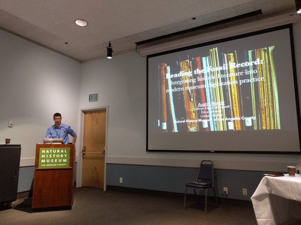man in blue shirt talking into a microphone behind a podium. Slides are visible on a screen behind him.