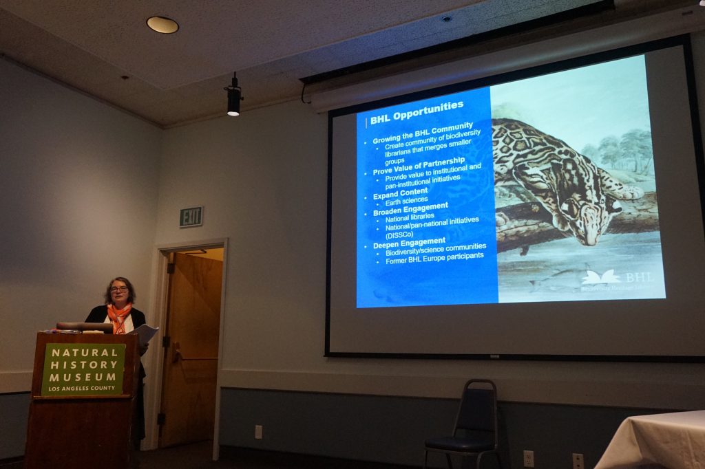 woman talking into a microphone behind a podium. Slides are visible on a screen behind her.