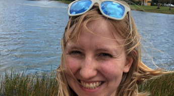 photo of a blonde woman with sunglasses on her head standing in front of a lake