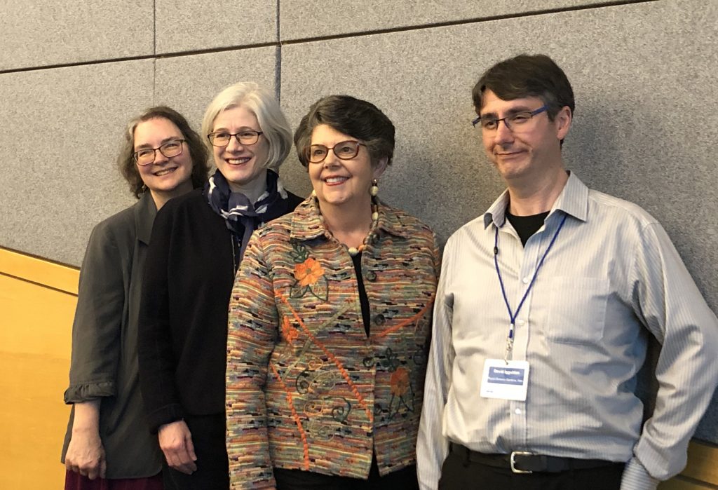 Group of three women and a man standing in front of a brown wall.