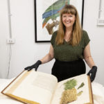 Woman with long brown hair, black clothes, and black gloves holds open a large book with scientific illustrations