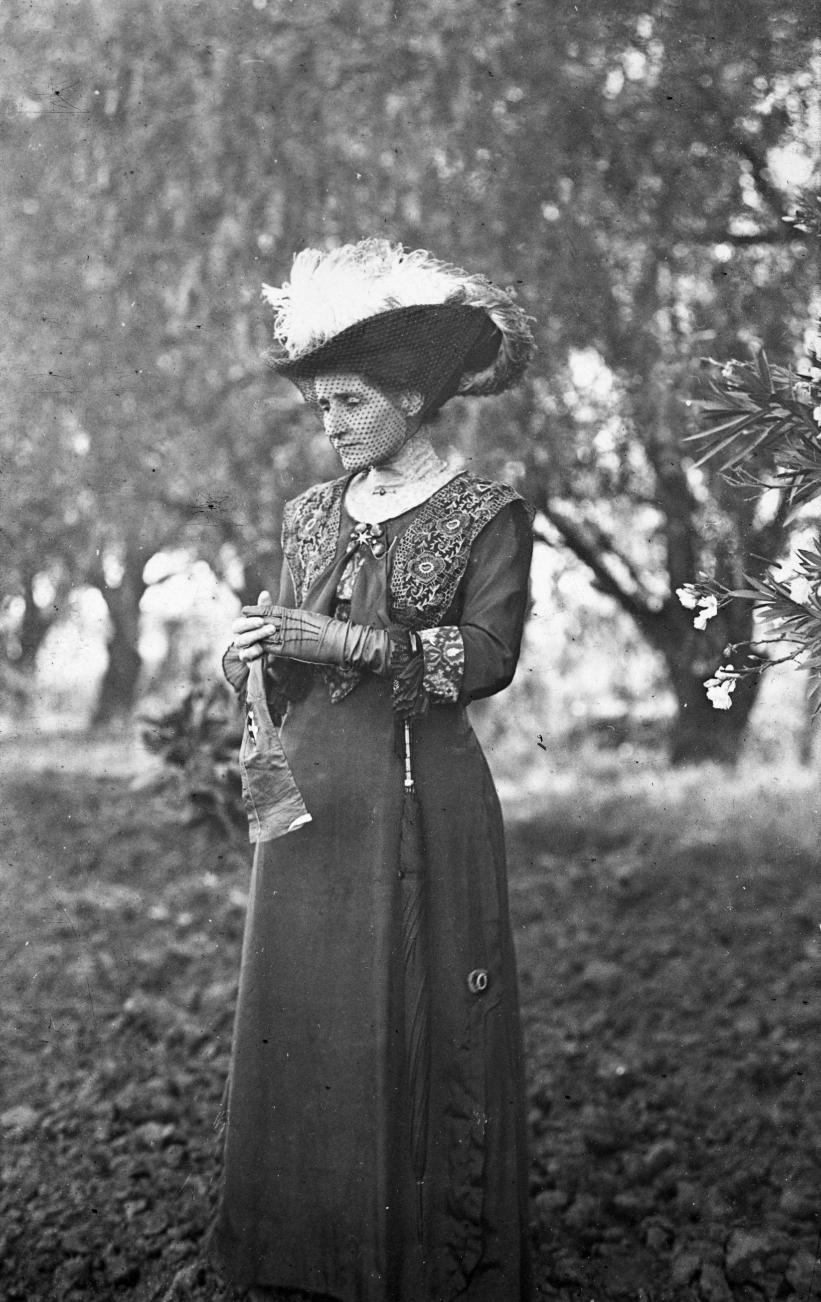 Black and white photograph of a woman in Victorian dress with an elaborate hat adorned with bird feathers. She stands in a forest.