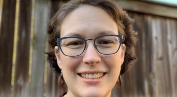 photograph of a human with glasses and brown hair standing in front of a fence outside.