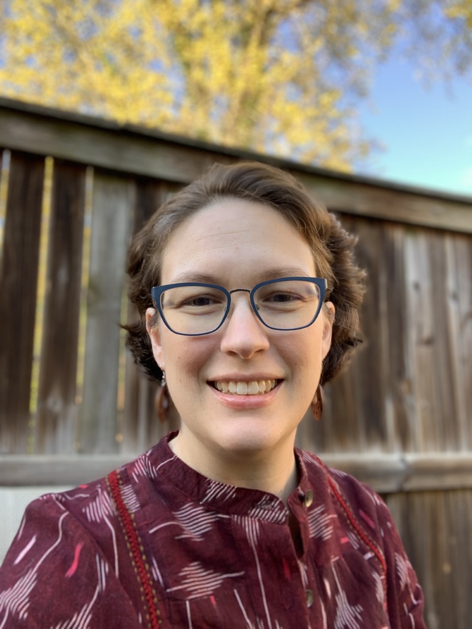 photograph of a human with glasses and brown hair standing in front of a fence outside.