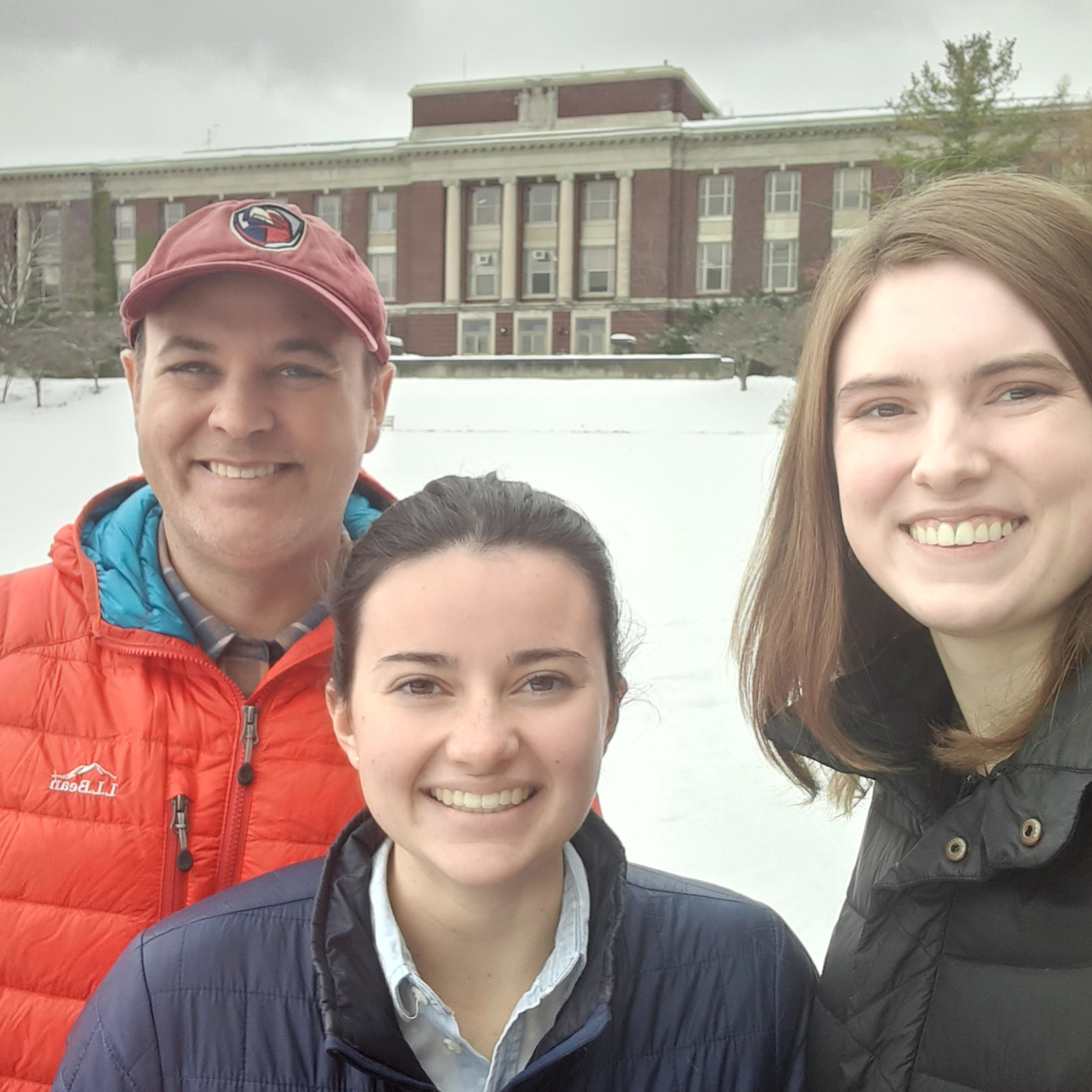 three people standing in the snow with a brick building in the back