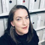 A woman with long dark hair and a black shirt stands in front of archival boxes