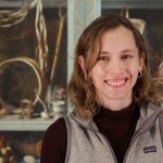 Woman with brown hair in a black shirt with tan vest standing in front of a museum case.