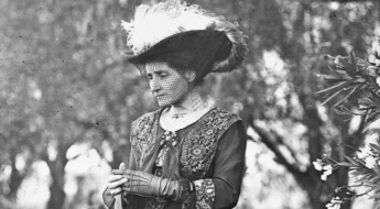 Black and white photo of a woman in a Victorian dress wearing an elaborate adorned with bird feathers. She stands in a forest.