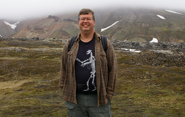 man in dark t-shirt with brown jacket standing in mossy landscape