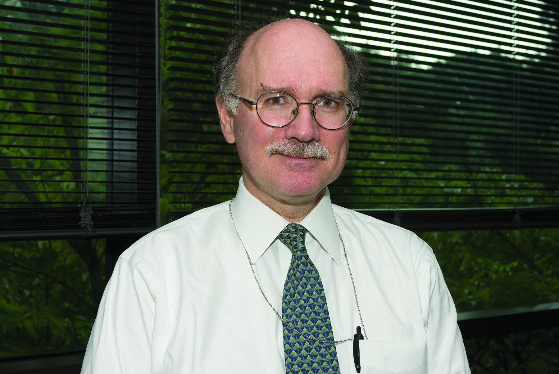 Caucasian male in a white button up shirt with a green tie. He smiles at the camera and wears glasses.
