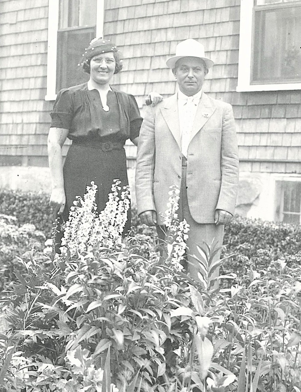 black and white photo of a woman and a man standing in a garden outside of a house.