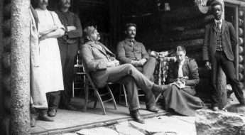 Black and white photo of a group of people standing on the porch of a log cabin.