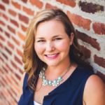 A blonde person in a blue shirt standing in front of a brick wall.