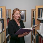 Person with red hair holding a book and standing in between book shelves.