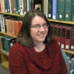 A person with brown hair and glasses in a red shirt sitting in front of a bookshelf