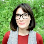 photograph of a human with glasses and brown hair standing in front of green foliage