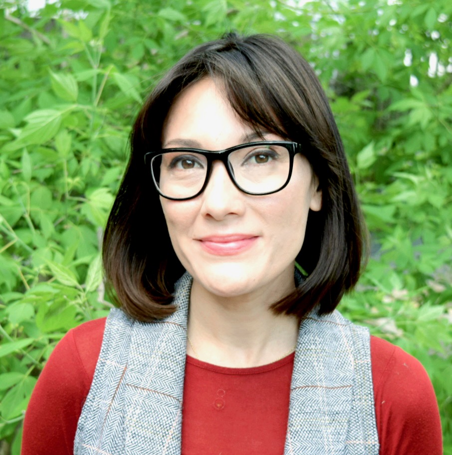 photograph of a human with glasses and brown hair standing in front of green foliage