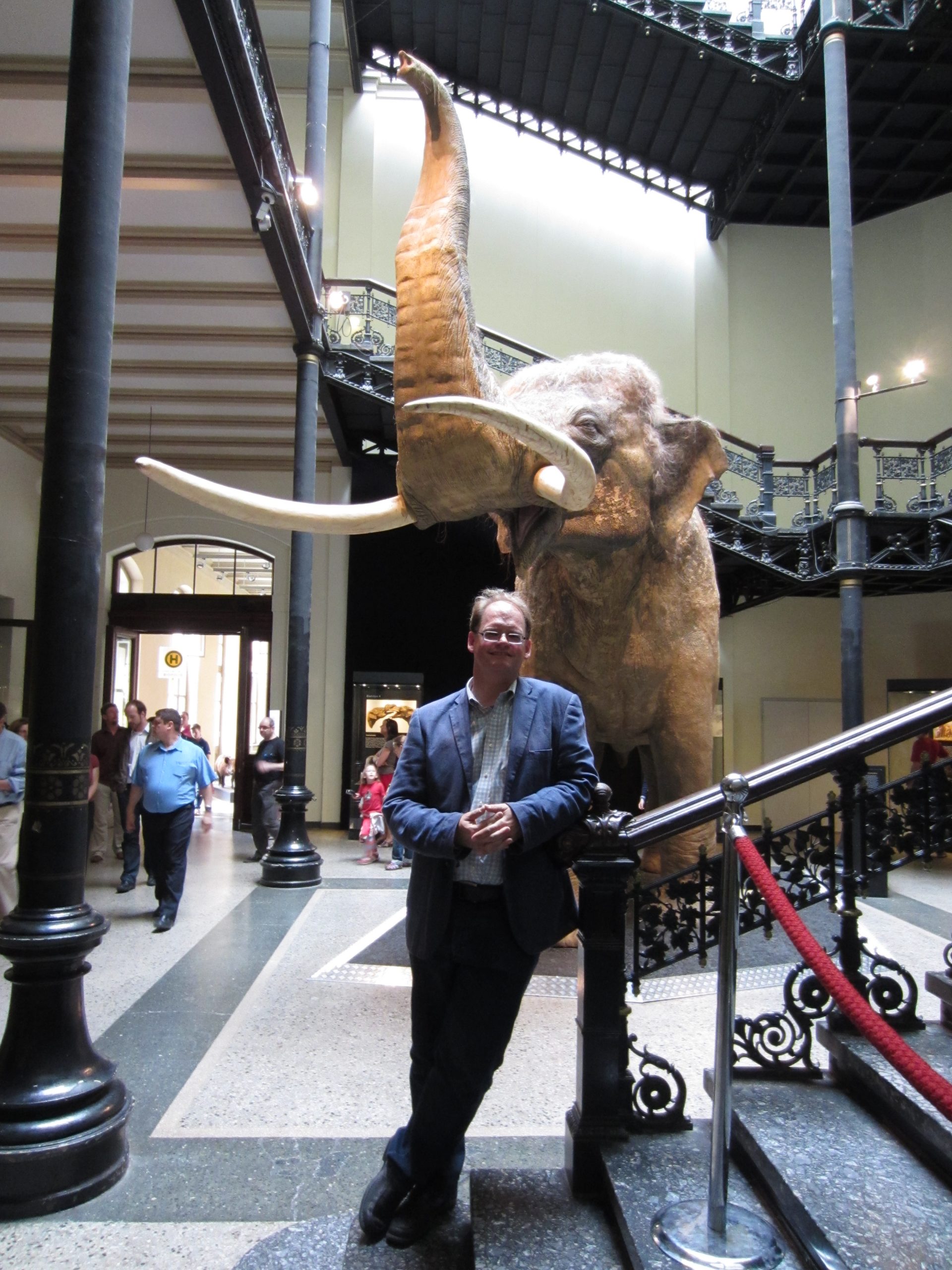 A man leans against a railing, posing in front of a life-sized museum display of an elephant