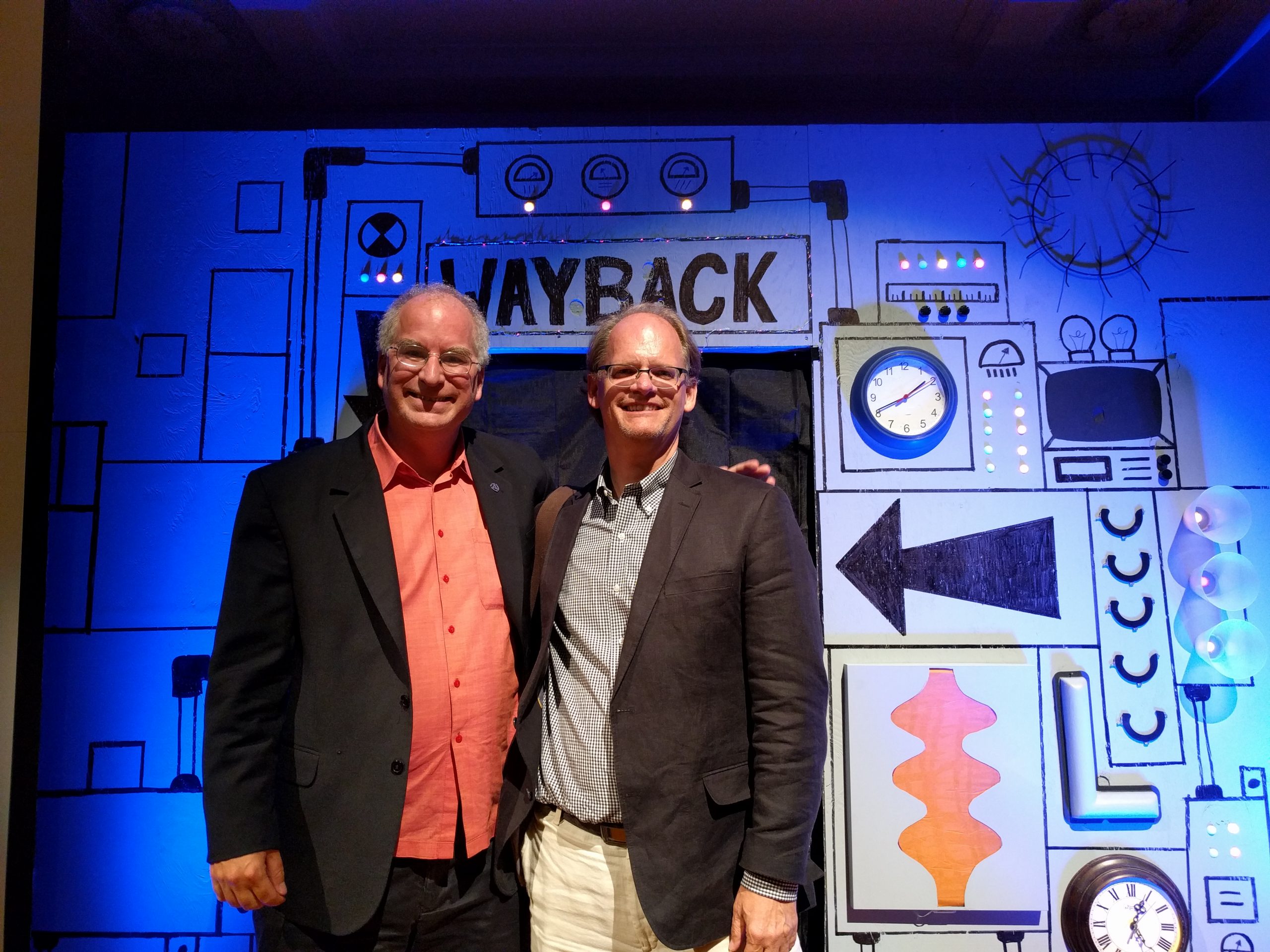 Two men in suit jackets and glasses smile and pose for a picture in front of a prop time machine