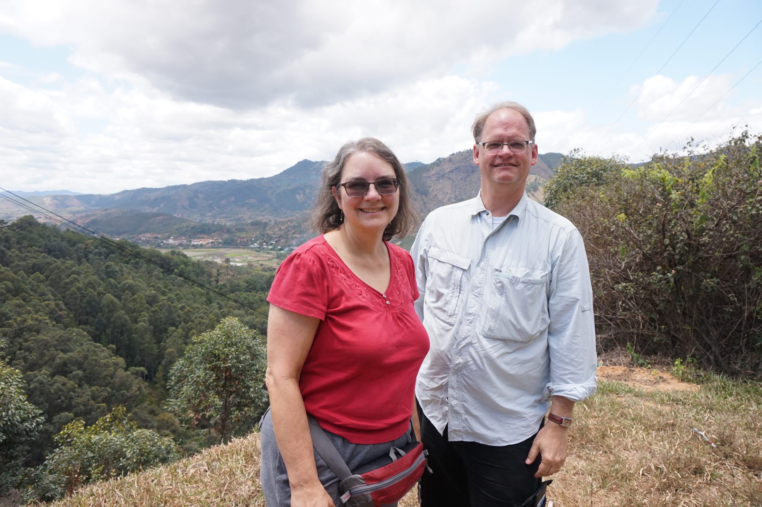 A woman in red shirt and sunglasses stands next to a man in a light blue shirt and glasses atop a hill overlooking a village