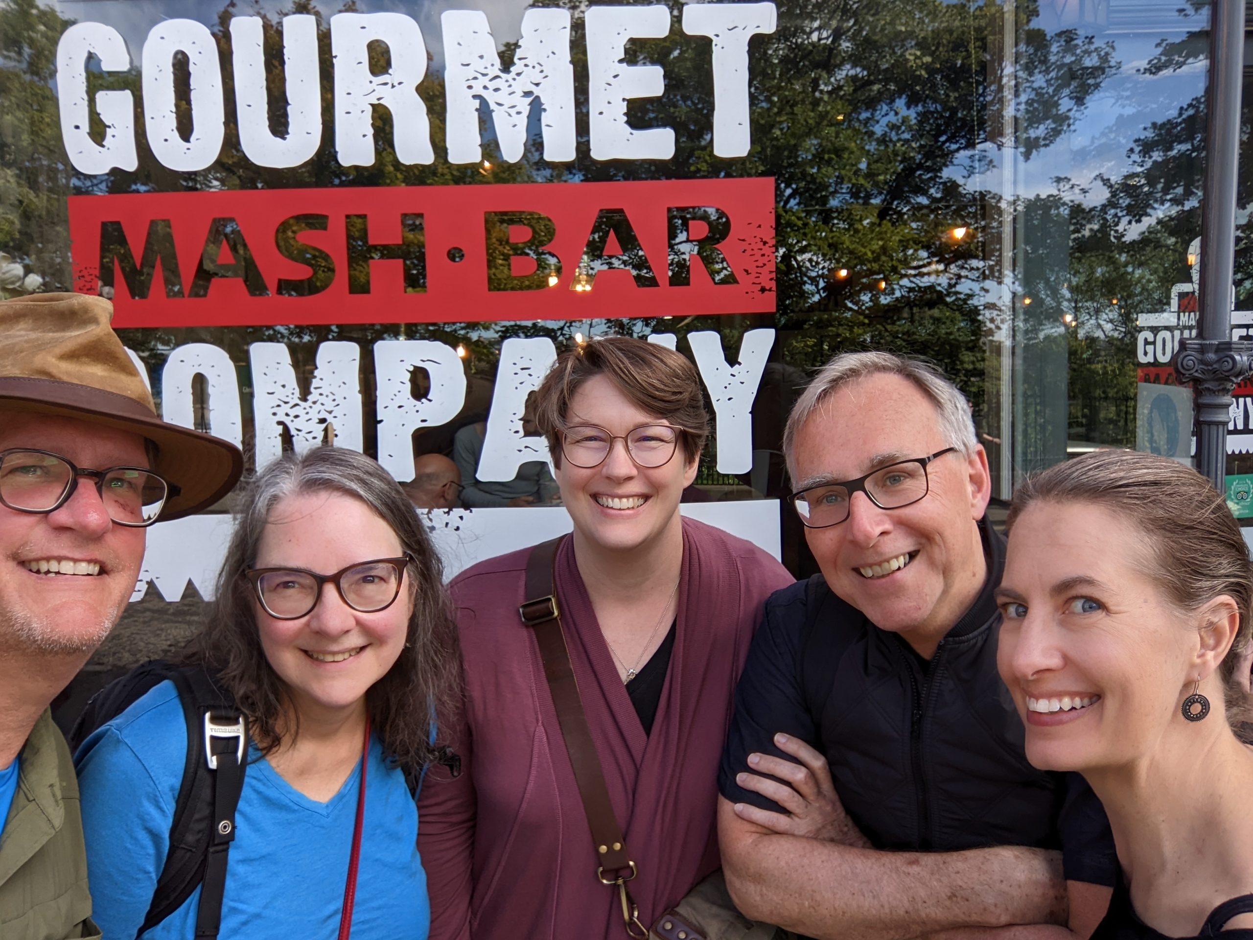 A group of five colleagues pose for a photo in front of a restaurant sign that reads Gourmet Mash Bar Company