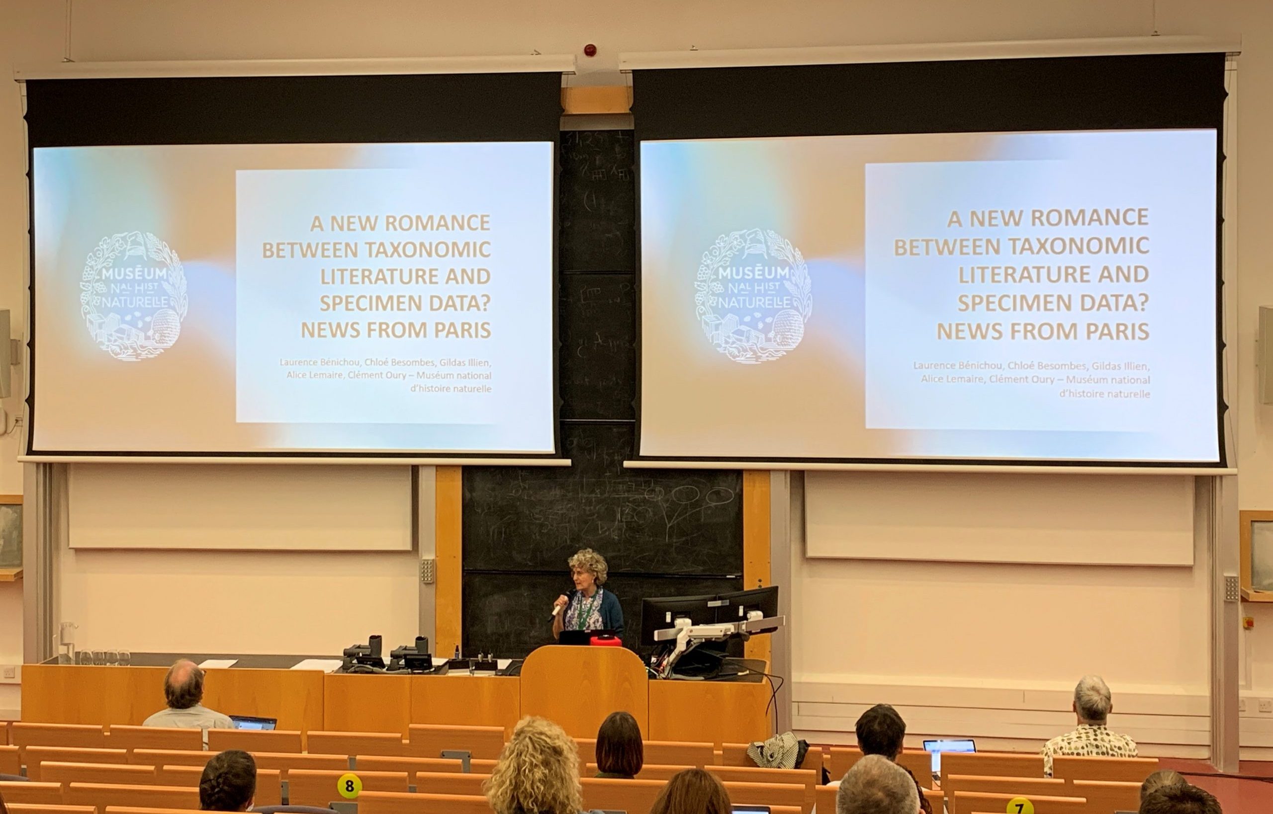 A speaker at a lectern presents to a lecture hall of attendees