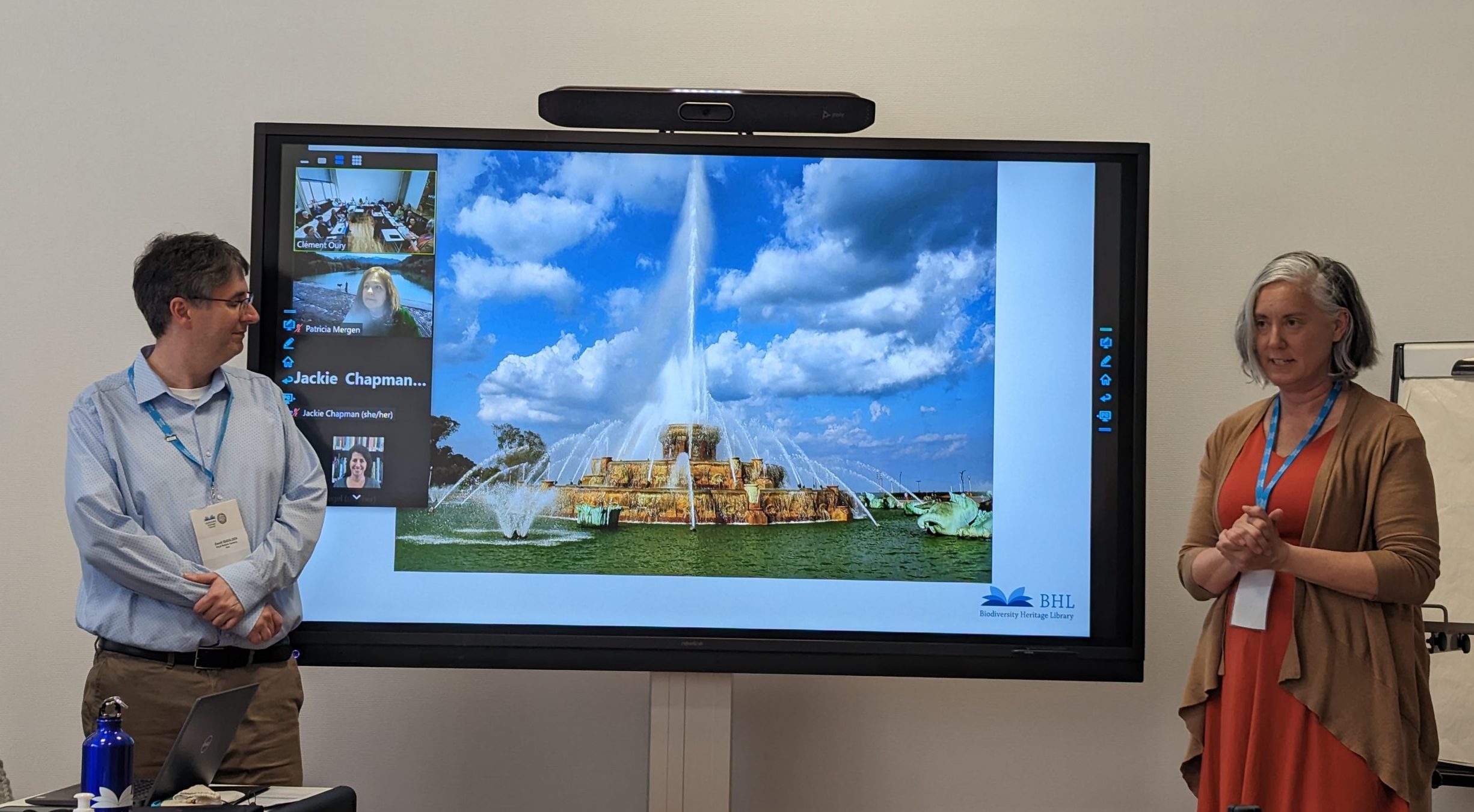 David Iggulden and Gretchen Rings stand next to a large digital display showcasing landmarks from Chicago including the Buckingham Fountain.