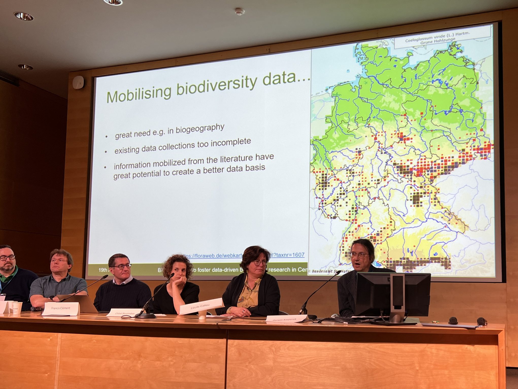 A panel of speakers on stage in front of a digital screen displaying biodiversity data