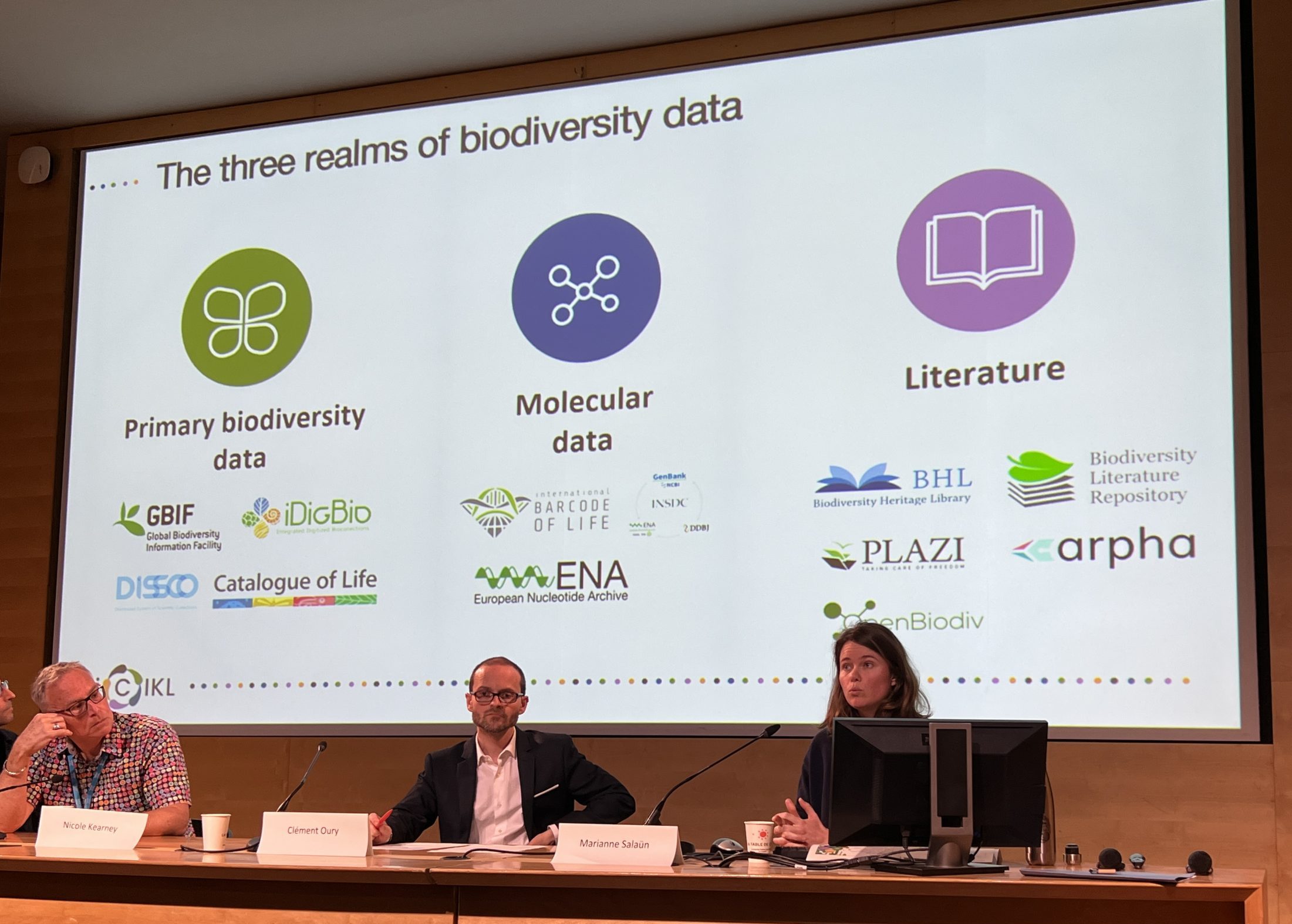 A panel of speakers on stage in front of a digital screen displaying the three realms biodiversity data - primary biodiversity data, molecular data, and literature
