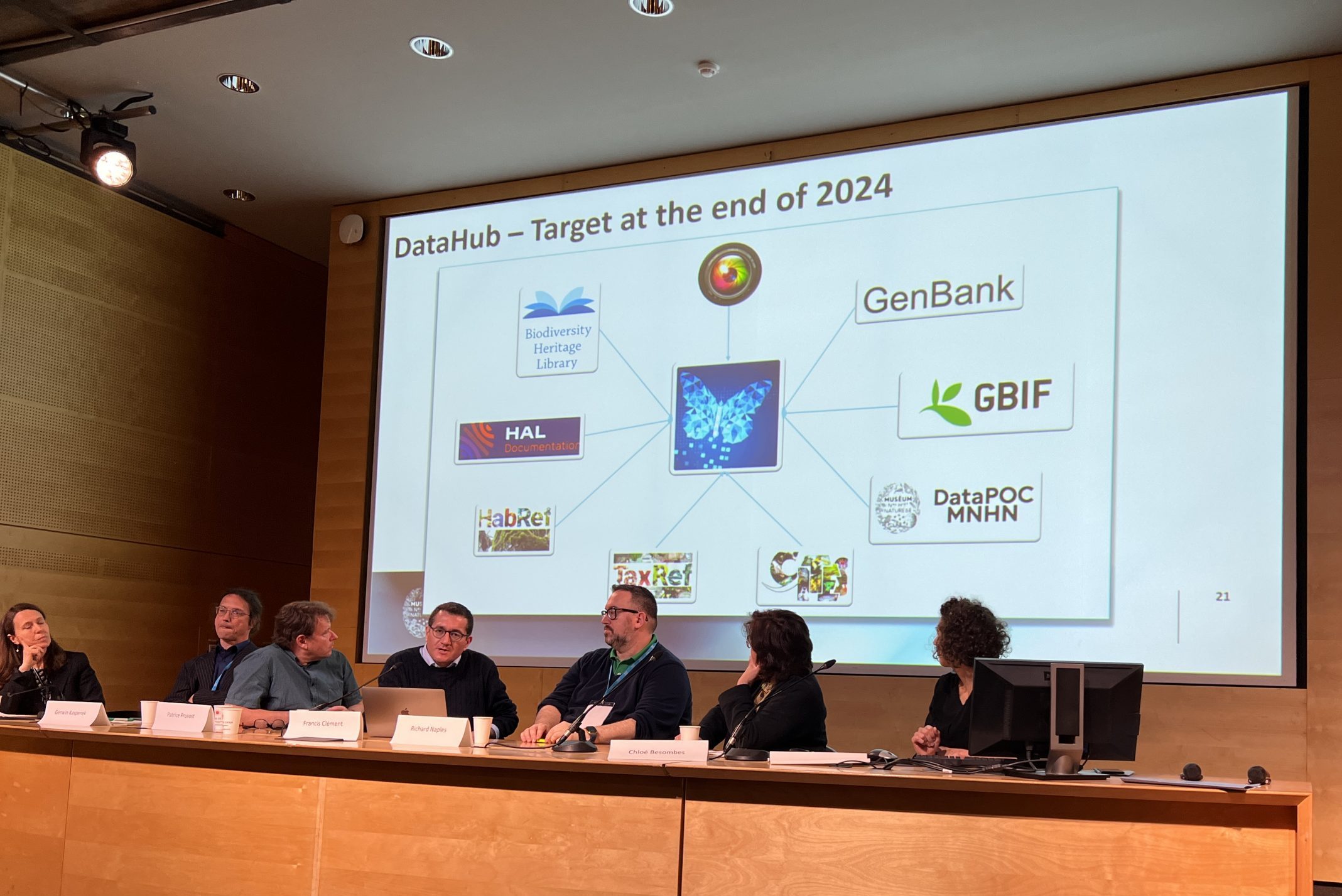 A panel of speakers present on stage in front of a digital display featuring the planned datahub for 2024