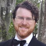 A man with brown hair and glasses, wearing a suit and bowtie standing in front of moss covered trees