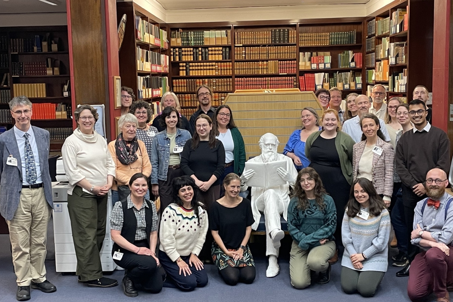 A group of 28 people posing for a group photo in a library around a life-sized white statue of Charles Darwin sitting with his legs crossed reading a book.