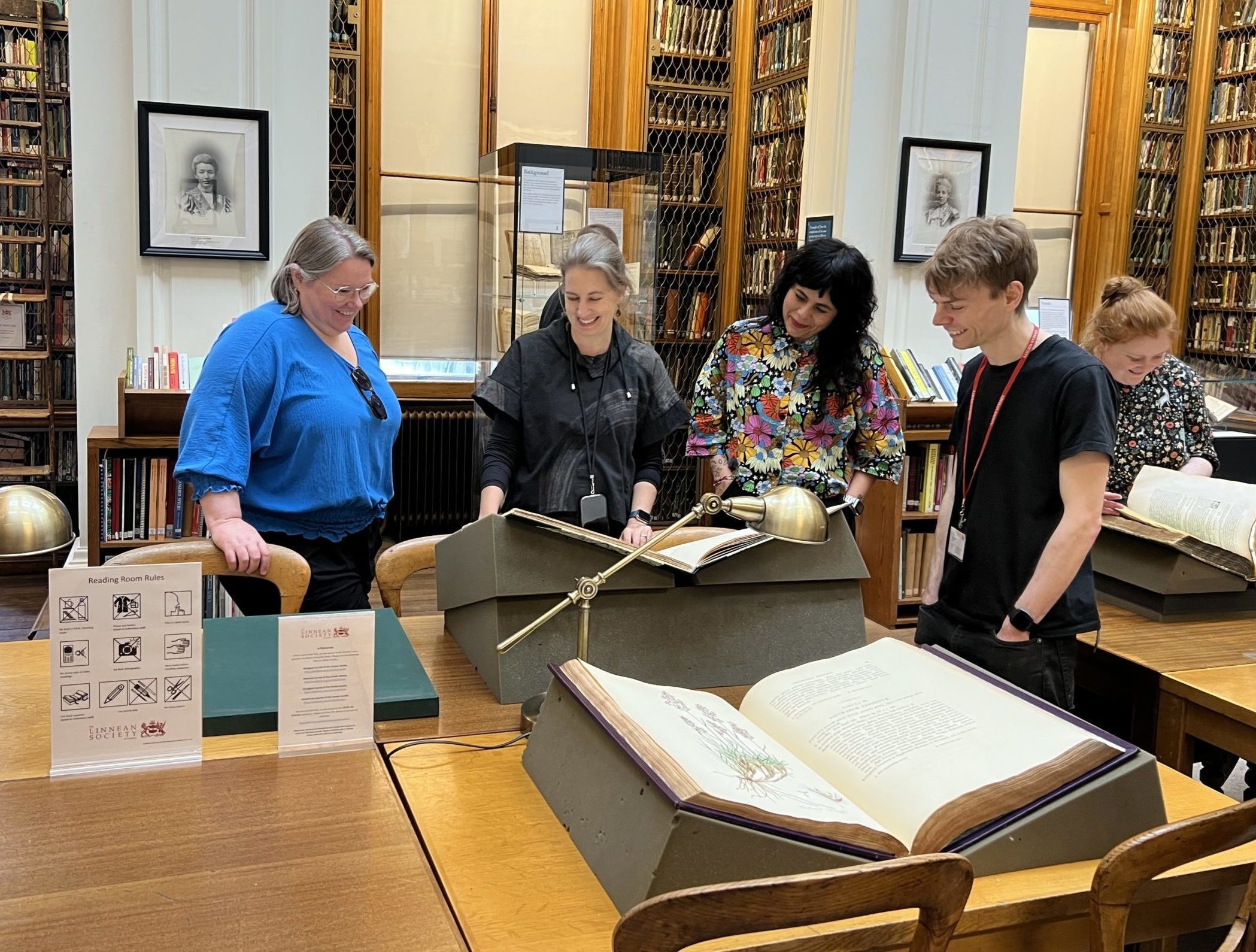 A group of smiling people looking at a large book open on a book stand on a table in a library.