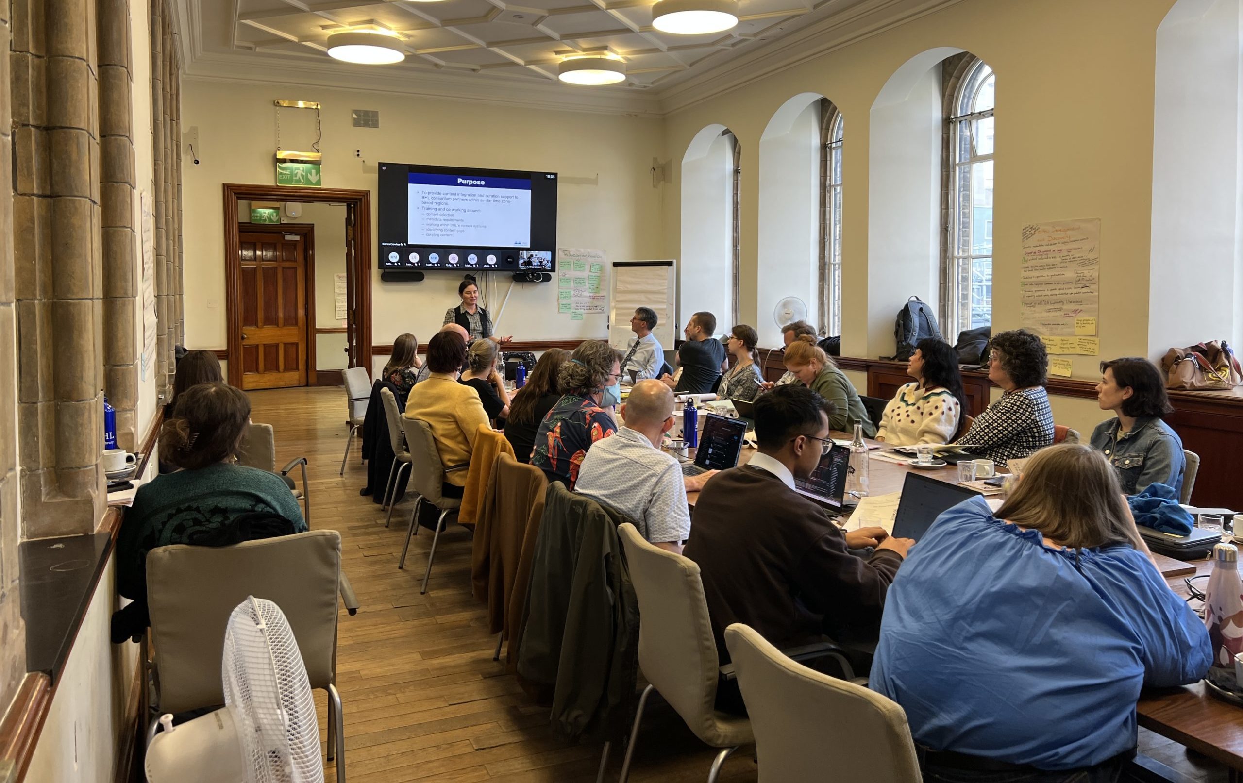 A group of people sitting at a long table watching a person give a presentation in front of a big screen.